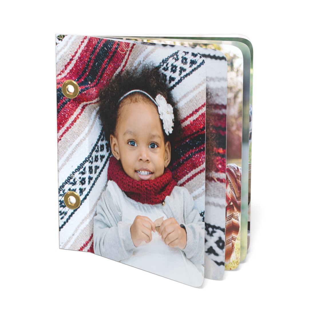 A small, open photo album with rounded corners, featuring a smiling child on the cover lying on a patterned blanket, with two gold grommets on the left edge and multiple photo pages fanning out on the right.