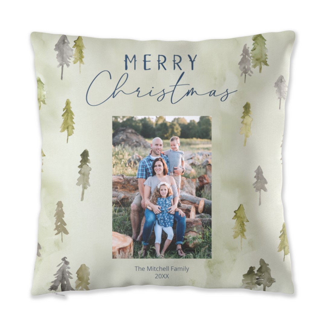 A square throw pillow with a light green background featuring watercolor evergreen trees, a central family photo of a man, woman, girl, and boy sitting on logs, and the text MERRY Christmas above the photo and The Mitchell Family 20XX below it.